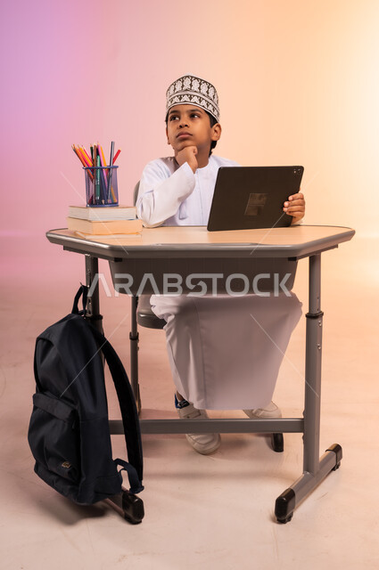طرائق تعليمية جديدة متطورة، بورتريه لطالب عربي خليجي عماني يرتدي الثوب التقليدي والقبعة العمانية يجلس في مقعده ويمسك بالجهاز اللوحي، النظر إلى الكاميرا بإيماءات السعادة والسرور، موسم العودة إلى المدارس، دمج التكنولوجيا بالتعليم في المدارس الحديثة، خلفية ملونة