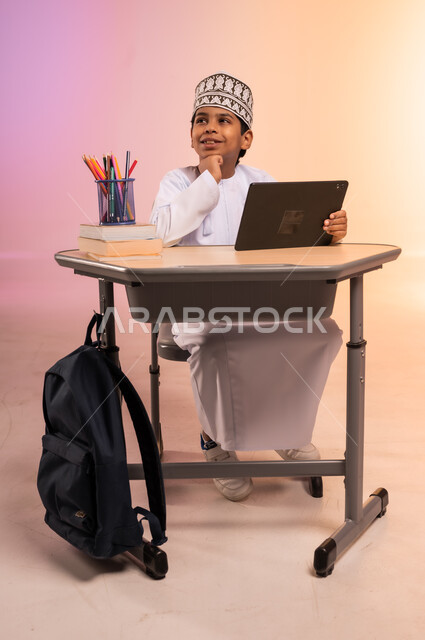 طرائق تعليمية جديدة متطورة، بورتريه لطالب عربي خليجي عماني يرتدي الثوب التقليدي والقبعة العمانية يجلس في مقعده ويمسك بالجهاز اللوحي، النظر إلى الكاميرا بإيماءات السعادة والسرور، موسم العودة إلى المدارس، دمج التكنولوجيا بالتعليم في المدارس الحديثة، خلفية ملونة