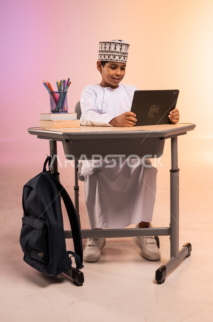 موسم العودة إلى المدارس، طرائق تعليمية جديدة متطورة، بورتريه لطالب عربي خليجي عماني يرتدي الثوب التقليدي والقبعة العمانية يجلس في مقعده يمسك بالجهاز اللوحي ويتفاعل مع الدروس عبر الانترنت، دمج التكنولوجيا بالتعليم في المدارس الحديثة، خلفية ملونة