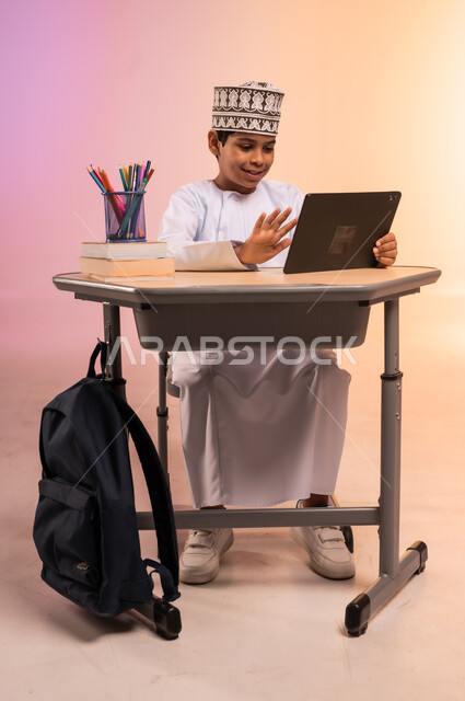 موسم العودة إلى المدارس، طرائق تعليمية جديدة متطورة، بورتريه لطالب عربي خليجي عماني يرتدي الثوب التقليدي والقبعة العمانية يجلس في مقعده يمسك بالجهاز اللوحي ويتفاعل مع الدروس عبر الانترنت، دمج التكنولوجيا بالتعليم في المدارس الحديثة، خلفية ملونة
