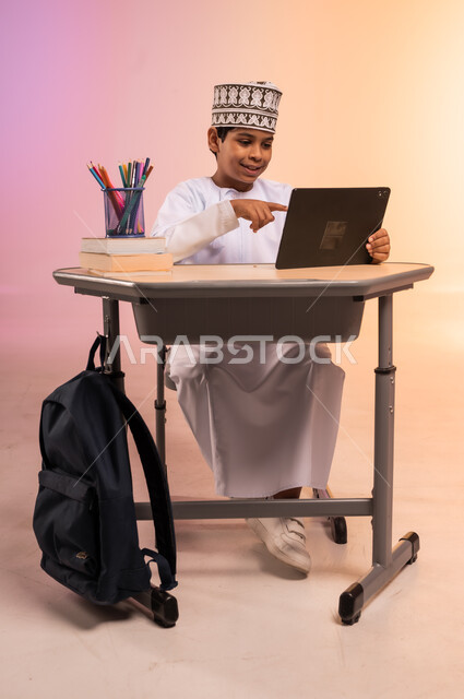 موسم العودة إلى المدارس، طرائق تعليمية جديدة متطورة، بورتريه لطالب عربي خليجي عماني يرتدي الثوب التقليدي والقبعة العمانية يجلس في مقعده يمسك بالجهاز اللوحي ويتفاعل مع الدروس عبر الانترنت، دمج التكنولوجيا بالتعليم في المدارس الحديثة، خلفية ملونة