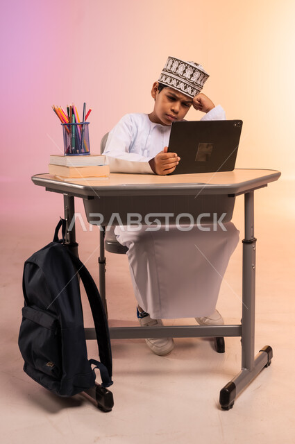 موسم العودة إلى المدارس، طرائق تعليمية جديدة متطورة، بورتريه لطالب عربي خليجي عماني يرتدي الثوب التقليدي والقبعة العمانية يجلس في مقعده يمسك بالجهاز اللوحي يتابع الدروس عبر الانترنت بإيماءات الملل، دمج التكنولوجيا بالتعليم في المدارس الحديثة، خلفية ملونة