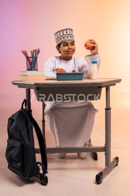 Back to school season, gestures of happiness and joy, school stationery and hanging black bag, portrait of an Arab Gulf Omani student wearing traditional dress and Omani hat sitting in his seat holding an apple from the lunch box in front of him, lunch box with healthy and nutritious snacks, colorful background