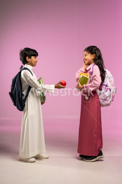 مشاركة الأصدقاء طعام الفطور, مفهوم العودة إلى المدارس, الاستعداد للعام الدراسي الجديد, بورتريه لولد عربي خليجي سعودي يرتدي الزي التقليدي يقوم بتقديم تفاحة حمراء لزميلته, الجسم بالطول الكامل, خلفية ملونة