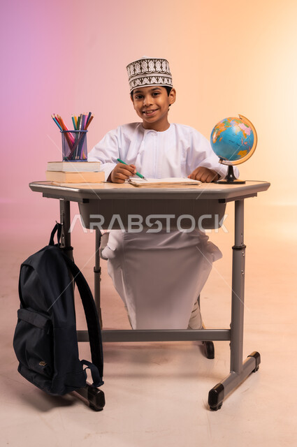 إيماءات السعادة والسرور بالعودة إلى المدارس، الجد والاجتهاد للوصول إلى النجاح والتفوق، بورتريه لطالب عربي خليجي عماني يرتدي الثوب التقليدي والقبعة العمانية يجلس في مقعده يمسك القلم بيده ويقوم بالكتابة والدراسة، قرطاسية مدرسية ومجسم للكرة الأرضية، خلفية ملونة