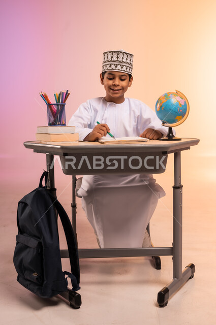 إيماءات السعادة والسرور بالعودة إلى المدارس، الجد والاجتهاد للوصول إلى النجاح والتفوق، بورتريه لطالب عربي خليجي عماني يرتدي الثوب التقليدي والقبعة العمانية يجلس في مقعده يمسك القلم بيده ويقوم بالكتابة والدراسة، قرطاسية مدرسية ومجسم للكرة الأرضية، خلفية ملونة