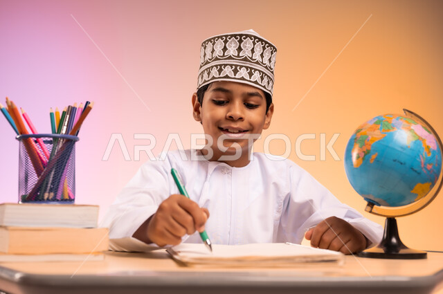 إيماءات السعادة والسرور بالعودة إلى المدارس، الجد والاجتهاد للوصول إلى النجاح والتفوق، بورتريه لطالب عربي خليجي عماني يرتدي الثوب التقليدي والقبعة العمانية يجلس في مقعده يمسك القلم بيده ويقوم بالكتابة والدراسة، قرطاسية مدرسية ومجسم للكرة الأرضية، خلفية ملونة
