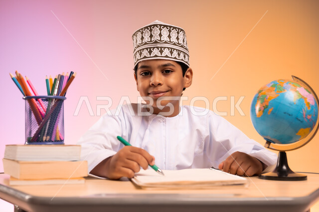 إيماءات السعادة والسرور بالعودة إلى المدارس، الجد والاجتهاد للوصول إلى النجاح والتفوق، بورتريه لطالب عربي خليجي عماني يرتدي الثوب التقليدي والقبعة العمانية يجلس في مقعده يمسك القلم بيده ويقوم بالكتابة والدراسة، قرطاسية مدرسية ومجسم للكرة الأرضية، خلفية ملونة