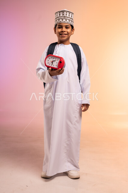 إيماءات النشاط والاستيقاظ باكراً، السعادة والسرور بالعودة إلى المدارس، الجد والاجتهاد للوصول إلى النجاح والتفوق، بورتريه لطالب عربي خليجي عماني يرتدي الثوب التقليدي والقبعة العمانية يحمل حقيبة الظهر المدرسية يقف ويمسك بساعة منبه أحمر اللون، خلفية ملونة