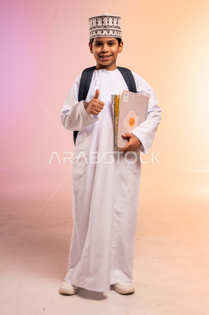 الجد والاجتهاد للوصول إلى النجاح والتفوق، موسم العودة إلى المدارس، بورتريه لطالب عربي خليجي عماني يرتدي الثوب التقليدي والقبعة يحمل حقيبة الظهر المدرسية يقف ويمسك بالكتب والمناهج العلمية، النظر للكاميرا بإيماءات السعادة والفرح، الجسم بالطول الكامل، خلفية ملونة