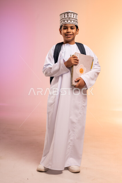 Hard work and diligence to achieve success and excellence, back to school season, portrait of an Arab Gulf Omani student wearing traditional dress and hat, carrying a school backpack, standing and holding books and scientific curricula, looking at the camera with gestures of happiness and joy, full body, colorful background