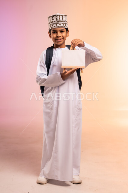 الجد والاجتهاد للوصول إلى النجاح والتفوق، موسم العودة إلى المدارس، بورتريه لطالب عربي خليجي عماني يرتدي الثوب التقليدي والقبعة يحمل حقيبة الظهر المدرسية يقف ويعرض لوحة أعلانية فارغة، النظر للكاميرا بإيماءات السعادة والفرح، الجسم بالطول الكامل، خلفية ملونة
