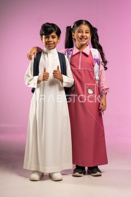 النظر إلى الكاميرا بإيماءات السعادة و السرور, الاستعداد للعام الدراسي الجديد, بورتريه لولد عربي خليجي سعودي يرتدي الزي التقليدي يقف بجانب زميلته و يرفع إصبعي الإبهام للأعلى, الجسم بالطول الكامل, خلفية ملونة