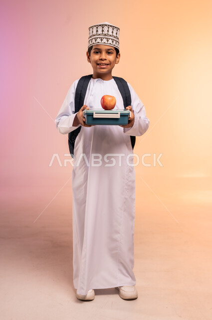 موسم العودة إلى المدارس، الجد والاجتهاد للوصول إلى النجاح والتفوق، بورتريه لطالب عربي خليجي عماني يرتدي الثوب التقليدي والقبعة يحمل حقيبة الظهر المدرسية يقف ويمسك علبة الطعام الصحي، النظر للكاميرا بإيماءات السعادة والفرح، الجسم بالطول الكامل، خلفية ملونة