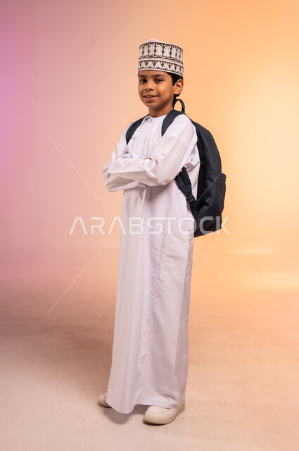 موسم العودة إلى المدارس، الجد والاجتهاد للوصول إلى النجاح والتفوق، بورتريه لطالب عربي خليجي عماني يرتدي الثوب التقليدي والقبعة يحمل حقيبة الظهر المدرسية يقف باستقامة ويدين متقاطعتين، النظر للكاميرا بإيماءات السعادة والفرح، الجسم بالطول الكامل، خلفية ملونة