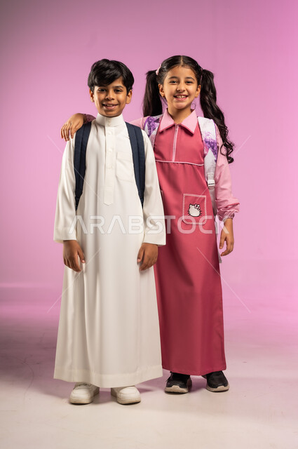 الاستعداد للعام الدراسي الجديد, مفهوم المحبة و المودة بين الزملاء, بورتريه لولد و بنت عربيان خليجيان سعوديان يرتديان الزي المدرسي ويحملان الحقائب المدرسية ينظران إلى الكاميرا بإيماءات السعادة و السرور, الجسم بالطول الكامل, خلفية ملونة