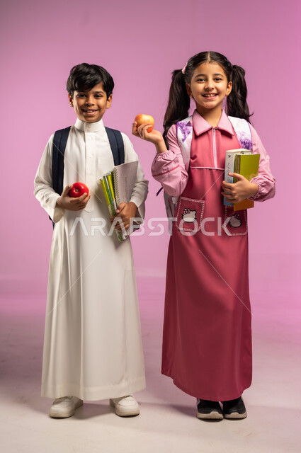 النظر إلى الكاميرا بإيماءات السعادة و السرور, مفهوم العودة إلى المدارس, الاستعداد للعام الدراسي الجديد, بورتريه لولد وبنت عربيان خليجيان سعوديان يرتديان الزي المدرسي و يحملان الكتب المدرسية و تفاحة حمراء, الجسم بالطول الكامل, خلفية ملونة
