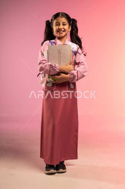 النظر إلى الكاميرا بإيماءات السعادة و السرور, الاستعداد للعام الدراسي الجديد, مفهوم العودة إلى المدارس, بورتريه لفتاة عربية خليجية سعودية ترتدي الزي المدرسي تحمل بيديها الكتب المدرسية, الجسم بالطول الكامل, خلفية ملونة