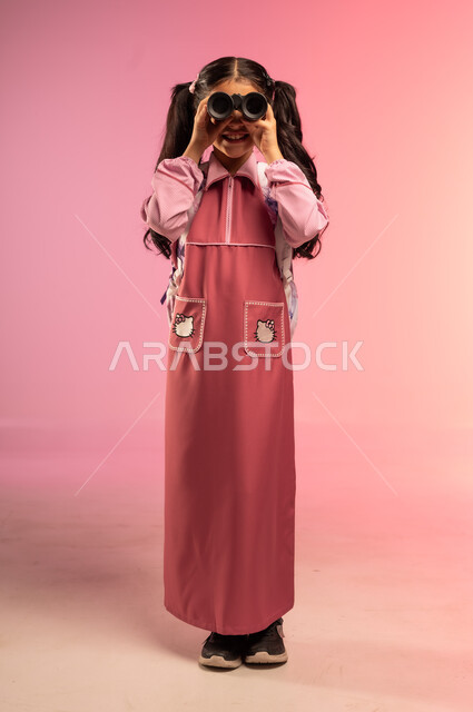 الاستعداد للعام الدراسي الجديد, مفهوم العودة إلى المدارس, التعليم في المملكة العربية السعودية, بورتريه لفتاة عربية خليجية سعودية ترتدي الزي المدرسي تنظر من خلال المنظار ذو العينين, الجسم بالطول الكامل, خلفية ملونة