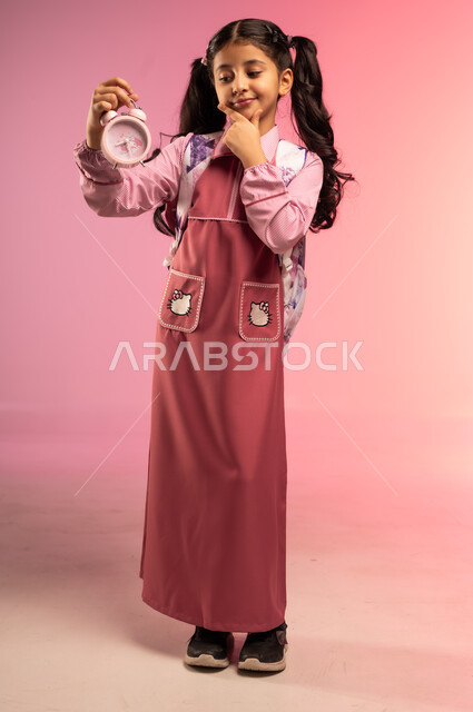 مفهوم تنظيم و إدارة الوقت, الاستعداد للعام الدراسي الجديد, التعليم في المملكة العربية السعودية, بورتريه لفتاة عربية خليجية سعودية ترتدي الزي المدرسي و تحمل على ظهرها حقيبة تمسك بيدها ساعة المنبه, الجسم بالطول الكامل, خلفية ملونة