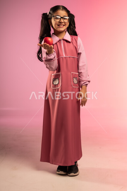 الفطور الصباحي الصحي و المفيد, الاستعداد للعام الدراسي الجديد, مفهوم العودة إلى المدارس, بورتريه لفتاة عربية خليجية سعودية مبتسمة ترتدي الزي المدرسي تنظر إلى الكاميرا و تحمل بيدها تفاحة حمراء, الجسم بالطول الكامل, خلفية ملونة