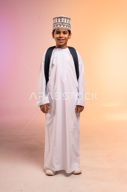 الوقوف باستقامة بتعابير الثقة بالنفس، موسم العودة إلى المدارس، الجد والاجتهاد للوصول إلى النجاح والتفوق، بورتريه لطالب عربي خليجي عماني يرتدي الثوب التقليدي والقبعة يحمل حقيبة الظهر المدرسية ينظر للكاميرا بإيماءات السعادة، الجسم بالطول الكامل، خلفية ملونة