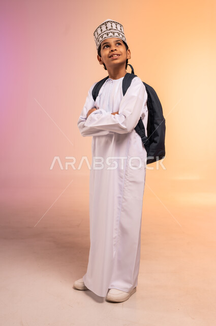الوقوف ياستقامة ويدين متقاطعتين، موسم العودة إلى المدارس، الجد والاجتهاد للوصول إلى النجاح والتفوق، بورتريه لطالب عربي خليجي عماني يرتدي الثوب التقليدي والقبعة يحمل حقيبة الظهر المدرسية ينظر للأعلى بإيماءات السعادة والفرح، الجسم بالطول الكامل، خلفية ملونة