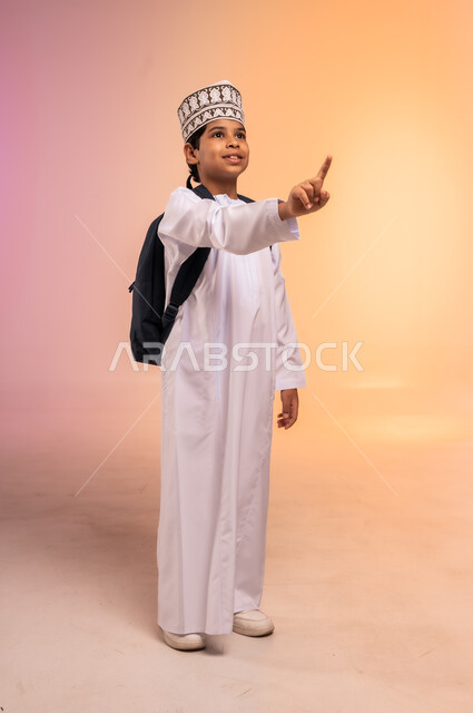 الإشارة إلى شيء ما بعيد، موسم العودة إلى المدارس، الجد والاجتهاد للوصول إلى النجاح والتفوق، بورتريه لطالب عربي خليجي عماني يرتدي الثوب التقليدي والقبعة يحمل حقيبة الظهر المدرسية ينظر للأعلى بإيماءات التركيز والاندماج، الجسم بالطول الكامل، خلفية ملونة