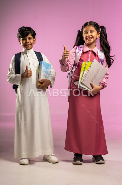 الإشارة بإصبع الإبهام للأعلى بتعابير الجودة, مفهوم العودة إلى المدارس, بورتريه لولد و بنت عربيان خليجيان سعوديان يرتديان الزي المدرسي يحملان الكتب و الأغراض المدرسية بيديهما, الجسم بالطول الكامل, خلفية ملونة