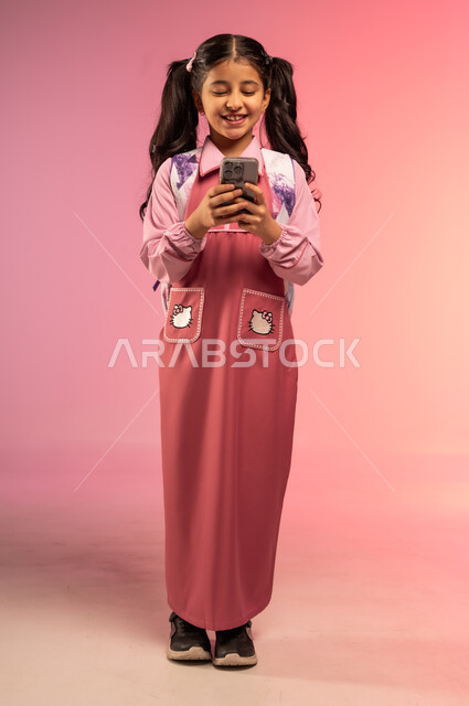 استخدام الأجهزة التقنية الحديثة في الحياة اليومية, الاستعداد للعام الدراسي الجديد, مفهوم العودة إلى المدارس, بورتريه لفتاة عربية خليجية سعودية ترتدي الزي المدرسي تقوم باستعمال الهاتف المحمول, الجسم بالطول الكامل, خلفية ملونة