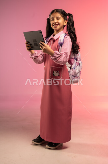 دمج الأجهزة التقنية الحديثة بالتعلم, الاستعداد للعام الدراسي الجديد, مفهوم العودة إلى المدارس, بورتريه لفتاة عربية خليجية سعودية ترتدي الزي المدرسي تحمل بيدها الجاهز اللوحي, الجسم بالطول الكامل, خلفية ملونة