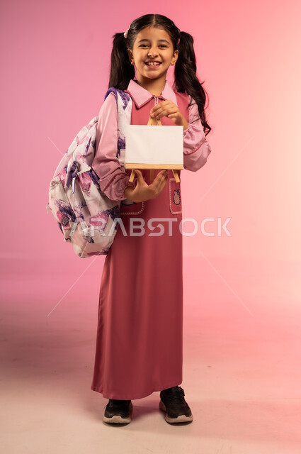 النظر إلى الكاميرا بإيماءات السعادة و السرور, الاستعداد للعودة إلى المدارس, بورتريه لفتاة عربية خليجية سعودية ترتدي الزي المدرسي تحمل بيدها لوحة بيضاء فارغة, الجسم بالطول الكامل, خلفية ملونة