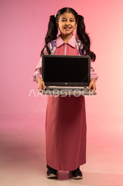 دمج الأجهزة التقنية الحديثة بالتعليم, الاستعداد للعام الدراسي الجديد, بورتريه لفتاة عربية خليجية سعودية ترتدي الزي المدرسي تحمل بيديها جهاز الحاسوب المحمول بشاشة سوداء فارغة, الجسم بالطول الكامل, خلفية ملونة