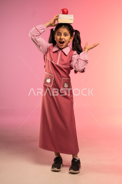 الاستعداد للعام الدراسي الجديد, حب التعلم و التعليم, النظر إلى الكاميرا بإيماءات السعادة و السرور, بورتريه لفتاة عربية خليجية سعودية ترتدي الزي المدرسي تضع على رأسها الكتب المدرسية وتفاحة حمراء, الجسم بالطول الكامل, خلفية ملونة