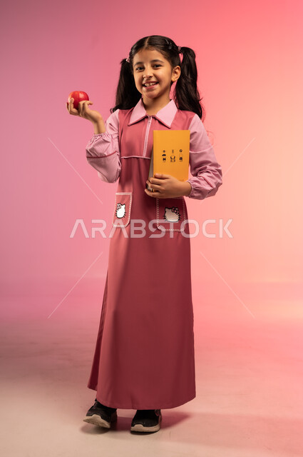 النظر إلى الكاميرا بإيماءات السعادة و السرور, الاستعداد للعام المدرسي الجديد, بورتريه لفتاة عربية خليجية سعودية ترتدي الزي المدرسي تحمل بيدها كتب مدرسية و تفاحة حمراء, الجسم بالطول الكامل, خلفية ملونة