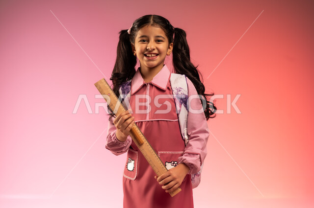الاستعداد للعام المدرسي الجديد, استخدام الأدوات المدرسية التعليمية, التعليم في المملكة العربية السعودية, بورتريه لفتاة عربية خليجية سعودية ترتدي الزي المدرسي تحمل بيدها مسطرة طويلة, خلفية ملونة