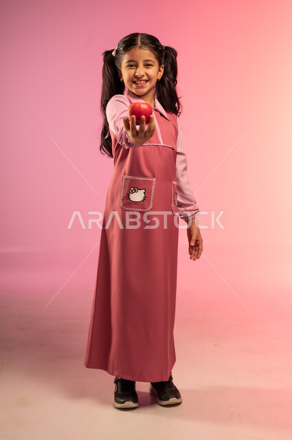 الاستعداد للعام الدراسي الجديد, النظر إلى الكاميرا بإيماءات السعادة و السرور, الفطور الصباحي الصحي و المفيد, بورتريه لفتاة عربية خليجية سعودية ترتدي الزي المدرسي تحمل بيدها تفاحة حمراء, الجسم بالطول الكامل, خلفية ملونة
