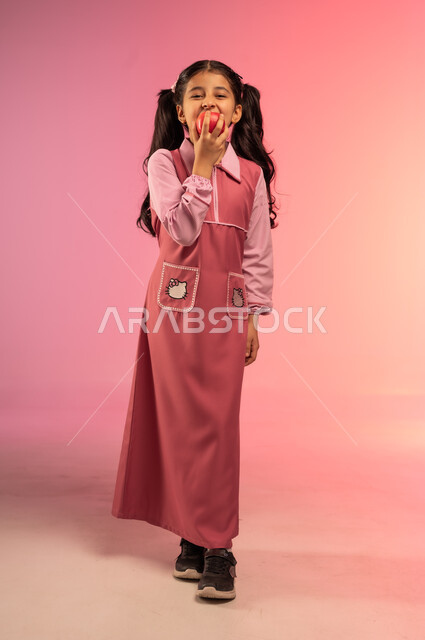 الفطور الصباحي الصحي و المفيد, الاستعداد للعام الدراسي الجديد, مفهوم العودة إلى المدارس, بورتريه لفتاة عربية خليجية سعودية ترتدي الزي المدرسي تأكل تفاحة حمراء, الجسم بالطول الكامل, خلفية ملونة