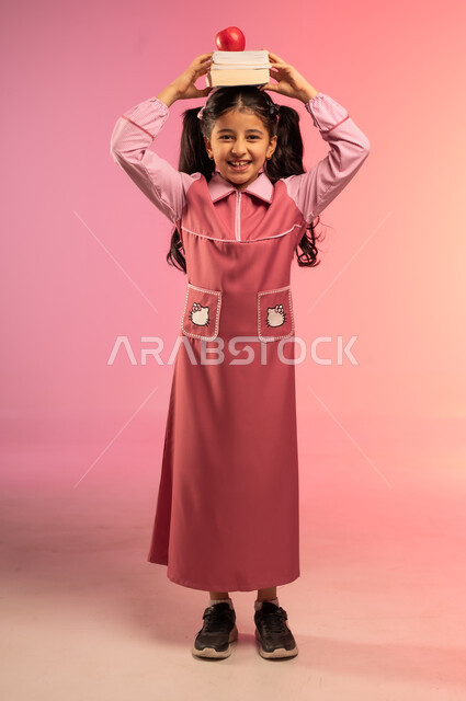 حب التعلم و التعليم, الاستعداد للعام الدراسي الجديد, النظر إلى الكاميرا بإيماءات السعادة و السرور, بورتريه لفتاة عربية خليجية سعودية ترتدي الزي المدرسي تضع على رأسها الكتب المدرسية وتفاحة حمراء, الجسم بالطول الكامل, خلفية ملونة