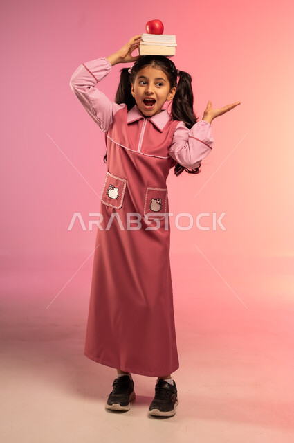 النظر إلى الكاميرا بإيماءات السعادة و السرور, حب التعلم و التعليم, الاستعداد للعام الدراسي الجديد, بورتريه لفتاة عربية خليجية سعودية ترتدي الزي المدرسي تضع على رأسها الكتب المدرسية وتفاحة حمراء, الجسم بالطول الكامل, خلفية ملونة