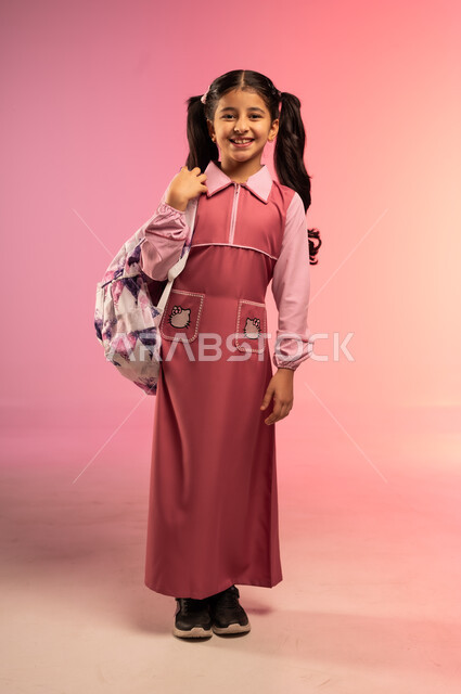 الاستعداد للعام الدراسي الجديد، التعليم في المملكة العربية السعودية, البهجة بالعودة إلى المدارس, بورتريه لفتاة عربية خليجية سعودية ترتدي الزي المدرسي تنظر إلى الكاميرا بإيماءات السعادة و السرور, الجسم بالطول الكامل, خلفية ملونة