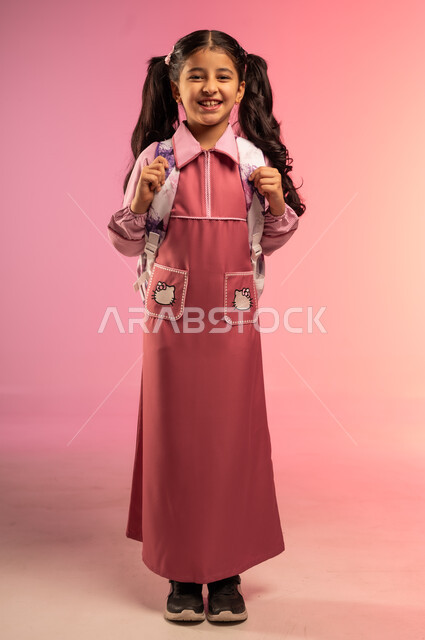 البهجة بالعودة إلى المدارس, الاستعداد للعام الدراسي الجديد، التعليم في المملكة العربية السعودية, بورتريه لفتاة عربية خليجية سعودية ترتدي الزي المدرسي تنظر إلى الكاميرا بإيماءات السعادة و السرور, الجسم بالطول الكامل, خلفية ملونة