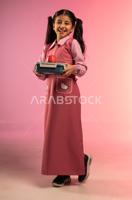 الاستعداد للعام الدراسي الجديد, النظر إلى الكاميرا بإيماءات السعادة و السرور, بورتريه لفتاة عربية خليجية سعودية ترتدي الزي المدرسي تحمل بيدها علبة طعام فوقها تفاحة حمراء, الجسم بالطول الكامل, خلفية ملونة