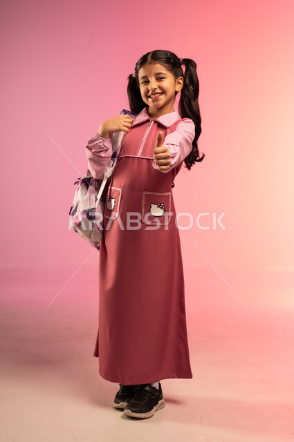الإشارة بإصبع الإبهام للأعلى بتعابير الجودة و الامتياز, البهجة بالعودة إلى المدارس, بورتريه لفتاة عربية خليجية سعودية ترتدي الزي المدرسي تنظر إلى الكاميرا بإيماءات السعادة و السرور, الجسم بالطول الكامل, خلفية ملونة