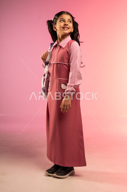 التحضير لبداية عام دراسي جديد, الاستعداد والتجهز للذهاب الى المدرسة, بورتريه لفتاة عربية خليجية سعودية ترتدي الزي المدرسي تحمل على ظهرها حقيبة مدرسية وتنظر إلى شيء ما بالأعلى, الجسم بالطول الكامل, خلفية ملونة