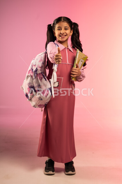 Thumbs up gesture with expressions of quality and excellence, joy of going back to school, portrait of a Saudi Arabian Gulf girl wearing school uniform looking at the camera with gestures of happiness and pleasure, full body, colorful background