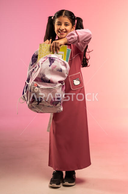 Joy of going back to school, getting ready and getting ready for the start of a new school year, portrait of a Saudi Arabian Gulf girl wearing a school uniform putting books and notebooks inside a school bag, full body, colorful background
