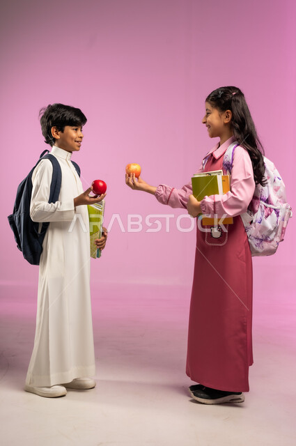 Friends sharing breakfast, back to school concept, getting ready for new school year, portrait of a Saudi Arabian Gulf boy and girl wearing school uniform and holding school books and a red apple, full body, colorful background