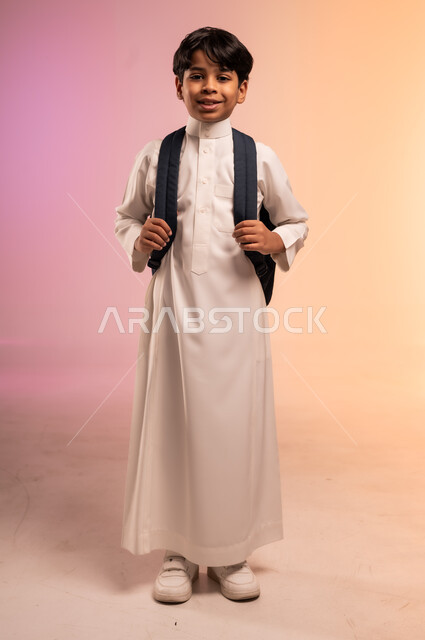 موسم العودة للمدارس في المملكة العربية السعودية، السعي نحو تحقيق حلم النجاح والتفوق، بورتريه لطالب عربي خليجي سعودي يرتدي الثوب التقليدي يحمل حقيبة الظهر المدرسية يقف باستقامة وينظر للكاميرا بإيماءات الثقة والبشاشة، الجسم بالطول الكامل، خلفية ملونة