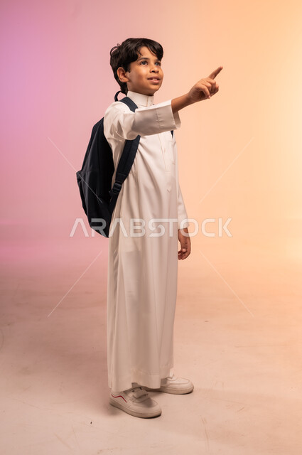رفع الاصبع والإشارة إلى شيء ما بعيد، موسم العودة للمدارس في السعودية، بورتريه لطالب عربي خليجي سعودي يرتدي الثوب التقليدي وحقيبة الظهر المدرسية يقف وينظر للأعلى بإيماءات الأمل وتحقيق الأحلام بالنجاح، الجسم بالطول الكامل، خلفية ملونة
